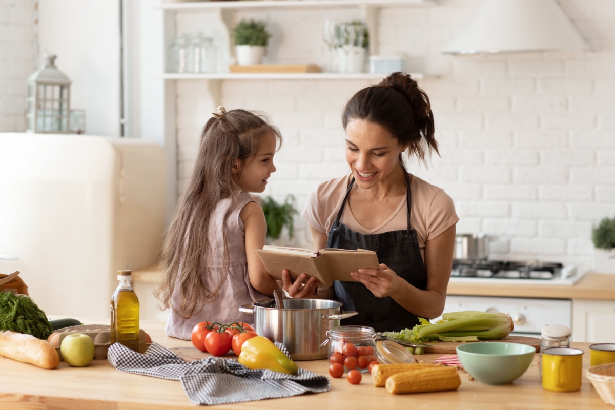 Mother reading cook book to daughter in kitchen. Photography by popcorner. Image via Shutterstock